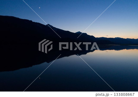 Depicting flat design dusk silhouette of mountain ridge reflecting on calm lake under crescent moon Depicting flat design dusk silhouette of mountain ridge reflecting on calm lake under crescent moon 133808667