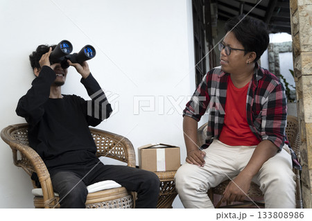 Two Indonesian southeast asian men sitting on a chair. Showing a package of a new pair of binoculars to his friend. Delivery service 133808986