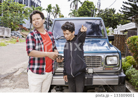 Two Indonesian southeast asian men in front of a blue car outside. One man is looking confused and asking the directions while the other is looking at a cellphone and pointing the way Two Indonesian southeast asian men in front of a blue car outside. One man is looking confused and asking the directions while the other is looking at a cellphone and pointing the way 133809001