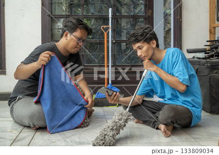 Two Indonesian southeast asian men sitting on the floor while holding cleaning equipment like a rag and a mop relaxed while looking at a cellphone screen. Cleaning home activity Two Indonesian southeast asian men sitting on the floor while holding cleaning equipment like a rag and a mop relaxed while looking at a cellphone screen. Cleaning home activity 133809034
