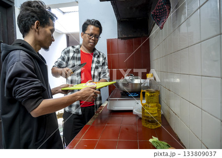 Two Indonesian southeast asian men inside a kitchen while preparing a meal together. Chopped ingredients on a chopping board 133809037