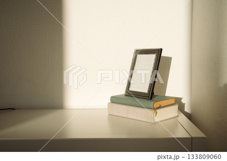 Empty silver photo frame resting on a stack of old books in direct sunlight. Casual home interior scene showing simple decor and vintage objects. Concept of home, interior, lifestyle, minimal, vintage 133809060
