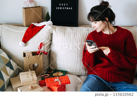 Young adult woman sits on a couch using a mobile phone surrounded by Christmas gifts and decorations. Mobile purchasing, virtual holiday shopping, conscious consumerism, remote connectivity. Young adult woman sits on a couch using a mobile phone surrounded by Christmas gifts and decorations. Mobile purchasing, virtual holiday shopping, conscious consumerism, remote connectivity. 133810562