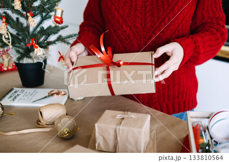 Young adult woman ties red ribbon on wrapped gift box on table with many eco-friendly supplies and tree. Plastic-free Christmas, zero-waste packaging, sustainable ribbons, green holiday design. 133810564