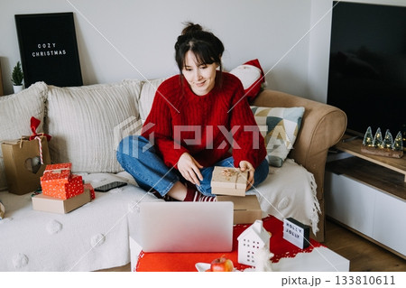 Young adult woman sits on sofa holding small wrapped gift box while using laptop for holiday shopping. Online shopping, seasonal ecommerce, Christmas sales, budget planning. 133810611