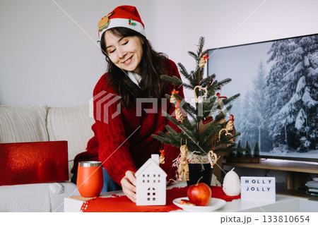 Smiling woman in a Santa hat arranges cozy holiday decorations next to Christmas tree with handmade ornaments. Nostalgic Christmas, handmade ornaments, natural decorations, warm seasonal tradition Smiling woman in a Santa hat arranges cozy holiday decorations next to Christmas tree with handmade ornaments. Nostalgic Christmas, handmade ornaments, natural decorations, warm seasonal tradition 133810654