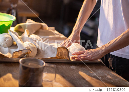 Hands of person placing raw bread dough loaves on proofing cloth, showing preparation for baking, with flour on wood surface, creating homemade bakery food Hands of person placing raw bread dough loaves on proofing cloth, showing preparation for baking, with flour on wood surface, creating homemade bakery food 133810803
