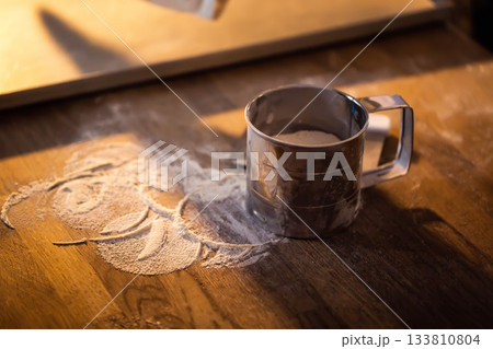Metal flour sifter on wooden surface with spilled white flour powder, preparing ingredients for baking recipe preparation, cooking process in kitchen setting with warm light Metal flour sifter on wooden surface with spilled white flour powder, preparing ingredients for baking recipe preparation, cooking process in kitchen setting with warm light 133810804