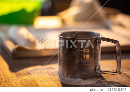 Metal flour sifter on wooden kitchen counter with flour. Baking ingredient preparation for bread or pastry making. Kitchen utensil close up on textured surface. Metal flour sifter on wooden kitchen counter with flour. Baking ingredient preparation for bread or pastry making. Kitchen utensil close up on textured surface. 133810805
