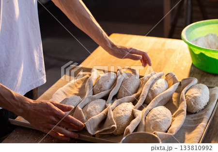 Bakers hands preparing raw bread dough loaves for baking process on a baking sheet, arranged in cloth for proofing, warm natural light illuminating kitchen scene Bakers hands preparing raw bread dough loaves for baking process on a baking sheet, arranged in cloth for proofing, warm natural light illuminating kitchen scene 133810992