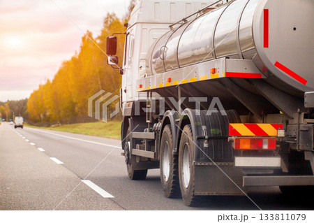 White tanker truck with large metallic tank trailer driving on asphalt highway, transportation of liquid cargo, journey during beautiful sunset with golden autumn trees lining roadside 133811079