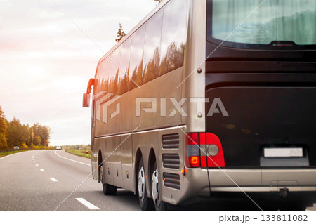 Large coach bus travels on asphalt road during sunset, warm sunlight reflects on vehicle side, trees line roadside, open sky on horizon, view of bus rear. 133811082