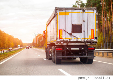 Rear view of white semi-trailer truck with yellow reflective stripes driving on multi-lane asphalt highway during golden hour, sun flare illuminating autumn forest lining roadside, distant vehicles 133811084