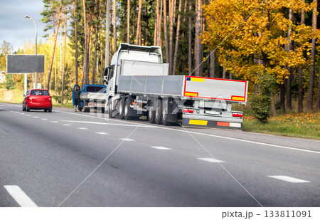 White semi-trailer truck with flatbed trailer parked roadside near red car and person on autumn highway alongside forest with yellow foliage White semi-trailer truck with flatbed trailer parked roadside near red car and person on autumn highway alongside forest with yellow foliage 133811091