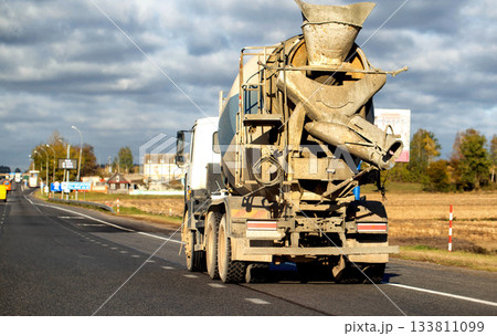 Cement mixer truck driving on asphalt highway road, rear view of vehicle showing large rotating drum and dirty surfaces, transportation of building materials under cloudy autumn sky 133811099