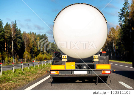 Rear view of tanker truck on highway, large white cylindrical tank for liquid transport, blue sky, forest alongside road, autumn trees, daytime outdoor scene Rear view of tanker truck on highway, large white cylindrical tank for liquid transport, blue sky, forest alongside road, autumn trees, daytime outdoor scene 133811103