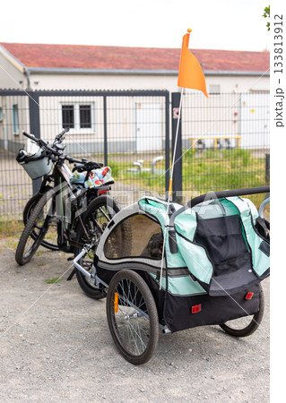 Parked bicycle attached child trailer stands on gravel near industrial fence, showing practical transport setup. Bright colors sturdy construction highlight mobility, family activity outdoor travel 133813912