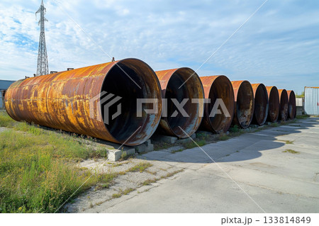 Large, rusted storage tanks are arranged in a row on a concrete path, surrounded by grass and a cloudy sky in the background Large, rusted storage tanks are arranged in a row on a concrete path, surrounded by grass and a cloudy sky in the background 133814849
