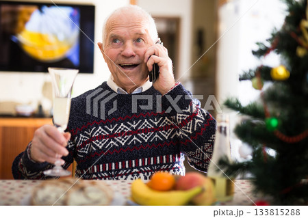 Elderly man wishes Merry Christmas by phone to friends and acquaintances sitting at the festive table in house Elderly man wishes Merry Christmas by phone to friends and acquaintances sitting at the festive table in house 133815288