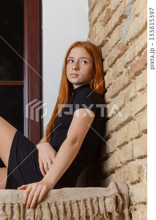 A young red-haired woman sits by a window with a brick wall A young red-haired woman sits by a window with a brick wall 133815397