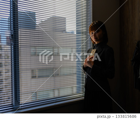 ブラインド越しの窓辺で何かを抱える若い女性 ブラインド越しの窓辺で何かを抱える若い女性 133815686