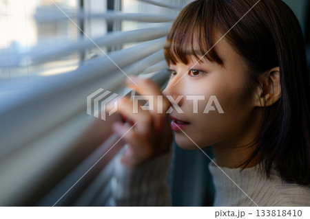 ブラインドの隙間から外を覗く若い女性のポートレート 133818410