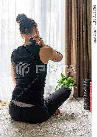 Young woman sitting on the floor at home using a massage ball to relieve neck pain. Self care routine for muscle tension and cervical spine health. Young woman sitting on the floor at home using a massage ball to relieve neck pain. Self care routine for muscle tension and cervical spine health. 133819768