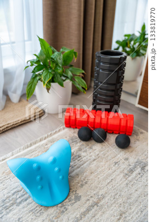Close-up of a blue cervical neck stretcher for spine decompression, placed on carpet at home. Foam rollers and massage balls visible in background, used for self-therapy and pain relief. 133819770