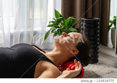 Young woman lying on the floor using a small red foam roller to massage her neck and occipital area. Daily self care routine to relieve muscle tension and improve cervical spine health. 133819775