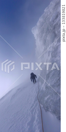 Climber trekking icy mountain ridge through dense fog. Solo mountaineer navigating a challenging snowy ridge against a foggy sky Climber trekking icy mountain ridge through dense fog. Solo mountaineer navigating a challenging snowy ridge against a foggy sky 133819821