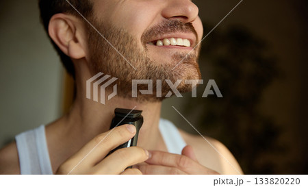 Smiling man trimming beard with electric grooming tool Smiling man trimming beard with electric grooming tool 133820220