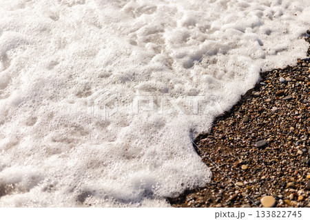 Gentle waves roll onto a sandy beach, creating a frothy foam along the shore. Small pebbles and grains of sand are visible as water recedes under the bright sun 133822745