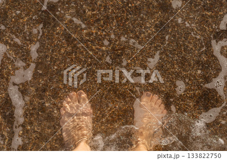 Bare feet stand in shallow water, feeling the gentle waves wash over them. Tiny pebbles and sand are visible under the clear water on a sunny afternoon Bare feet stand in shallow water, feeling the gentle waves wash over them. Tiny pebbles and sand are visible under the clear water on a sunny afternoon 133822750