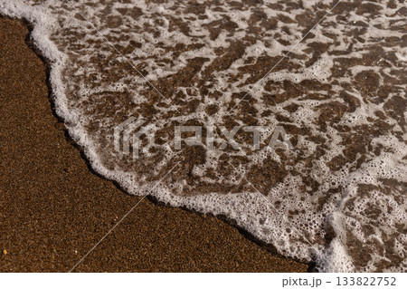 Soft waves are lapping against the golden sand on a beach during a bright day. The water creates a frothy edge as it retreats back into the sea 133822752
