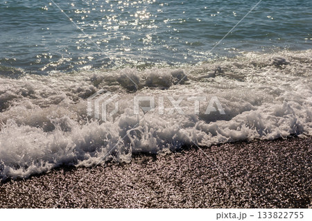 Waves roll onto a sandy beach under bright sunlight, creating a sparkling effect on the water. The tranquil scene invites relaxation and enjoyment of nature's beauty 133822755