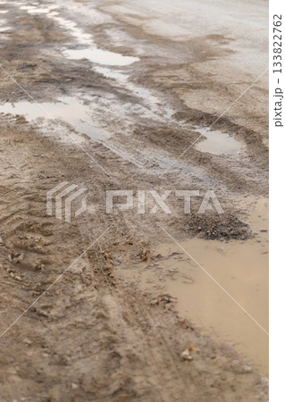 A muddy dirt road stretches across the landscape, with visible tire tracks and puddles forming after a recent rainfall. The scene captures a typical rural environment A muddy dirt road stretches across the landscape, with visible tire tracks and puddles forming after a recent rainfall. The scene captures a typical rural environment 133822762