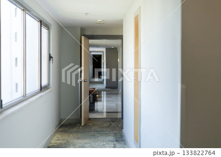 A spacious hotel corridor shows an open door revealing a cozy waiting area. Natural light fills the hallway, enhancing its modern and inviting atmosphere A spacious hotel corridor shows an open door revealing a cozy waiting area. Natural light fills the hallway, enhancing its modern and inviting atmosphere 133822764
