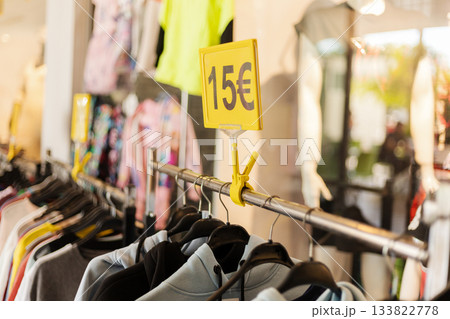 A row of clothing hangs neatly on hangers in a retail store. A bright yellow tag shows the price of 15 euros, highlighting a sale. The environment is well-lit with colorful garments A row of clothing hangs neatly on hangers in a retail store. A bright yellow tag shows the price of 15 euros, highlighting a sale. The environment is well-lit with colorful garments 133822778