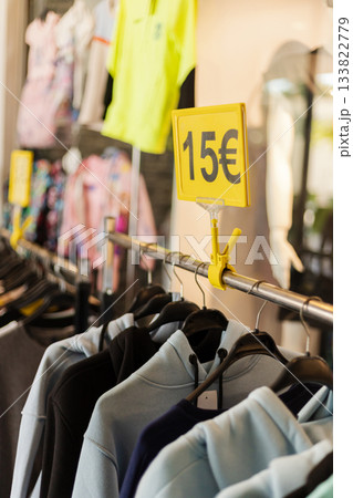 In a retail store, clothes hang on a metal rack with a bright yellow sign displaying a price of 15 euros, inviting customers to shop for affordable fashion 133822779