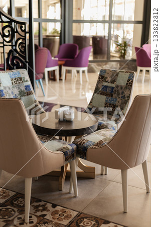 A cozy seating area features two unique chairs and a round table. Bright natural light fills the space, creating a welcoming ambiance for visitors to relax or socialize 133822812