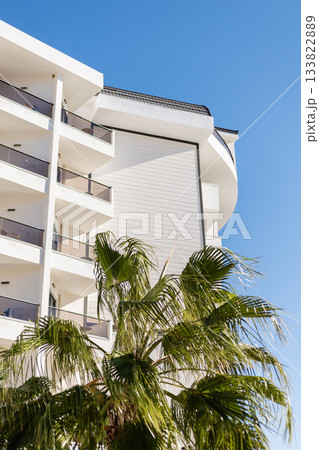 A contemporary building features multiple balconies and a palm tree in the foreground. The clear blue sky highlights the sunny atmosphere of this pleasant location 133822889