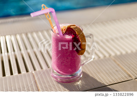 A vibrant pink drink is served in a clear glass, topped with a slice of tropical fruit and a straw. Sunlight shines down on the poolside, creating a perfect summertime vibe 133822896