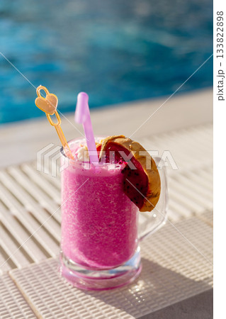 A vibrant pink beverage garnished with a slice of dragon fruit and pineapple rests on the edge of a pool, capturing a relaxing summer vibe 133822898