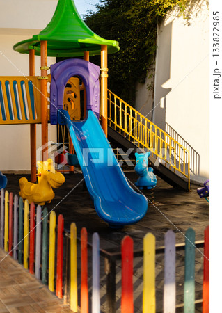 A vibrant playground features a blue slide, playful animal figures, and colorful fencing. The sunny setting invites children to engage in outdoor fun and play 133822985