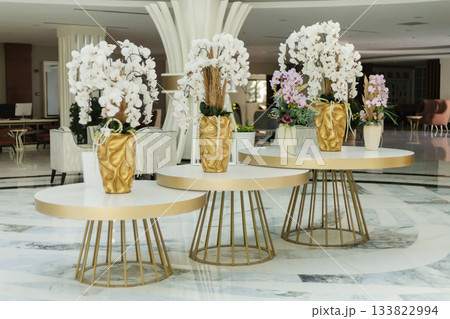 Three stylish tables are displayed in a hotel lobby, adorned with beautiful floral arrangements in decorative vases. The setting features modern decor and bright lighting 133822994