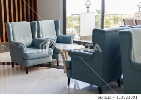 Two stylish blue armchairs sit next to a small round table, creating a cozy seating area. Large windows let in natural light, enhancing the modern decor 133823001