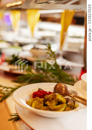 A bowl of mixed vegetables sits at the front of a buffet line in a restaurant. Fresh greens, red peppers, and assorted options fill the background, offering a variety of choices A bowl of mixed vegetables sits at the front of a buffet line in a restaurant. Fresh greens, red peppers, and assorted options fill the background, offering a variety of choices 133823028