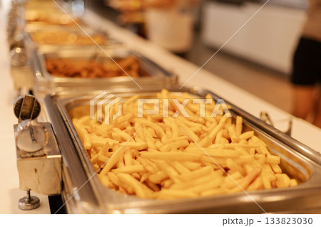 A buffet features trays filled with golden, crispy french fries. Diners are enjoying a hearty lunch surrounded by various food options. The setting is bright and inviting 133823030