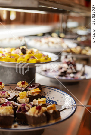 An enticing spread of colorful desserts is arranged on plates at a lively event. Guests enjoy a variety of sweets including chocolate cakes and fruit tarts. The atmosphere is joyful and inviting An enticing spread of colorful desserts is arranged on plates at a lively event. Guests enjoy a variety of sweets including chocolate cakes and fruit tarts. The atmosphere is joyful and inviting 133823037