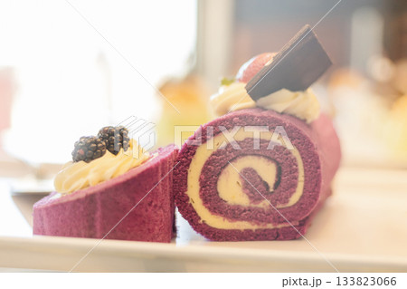 A beautifully arranged purple roll cake sits on a white plate, garnished with whipped cream, chocolate, and fresh blackberries. The cake showcases an inviting swirl design A beautifully arranged purple roll cake sits on a white plate, garnished with whipped cream, chocolate, and fresh blackberries. The cake showcases an inviting swirl design 133823066